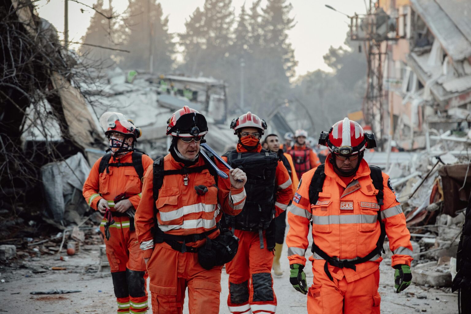 Men in orange walking through wreckage of an earthquakes aftermath.