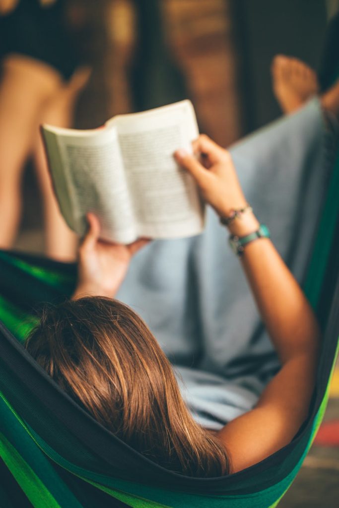 Person with light brown hair reading a book in a green hammock.