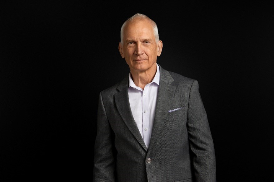 Don standing in a grey suit with black background.